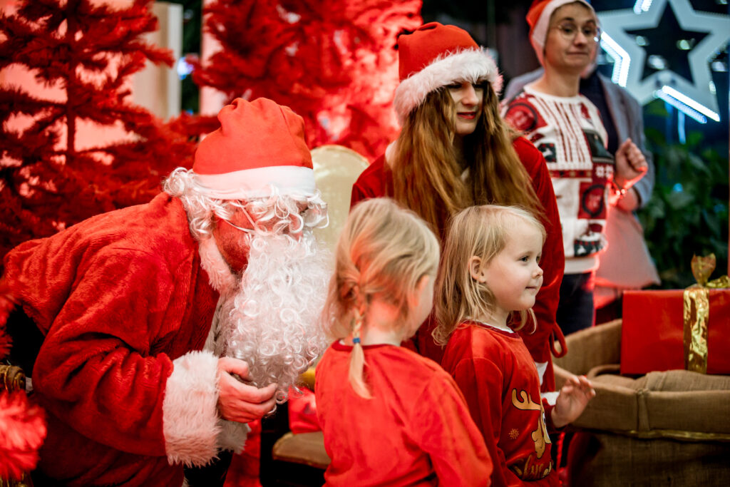 Zdjęcie. Mikołajki w KCK. Mikołaj pochyla się w kierunku dwoch małych dziewczynek ubranych w czerwone bluzki.