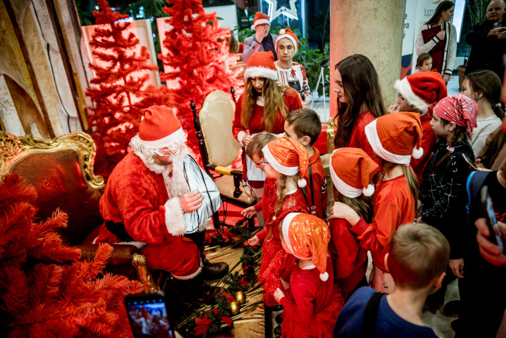 Zdjęcie. Mikołajki w KCK. Mikołaj obstąpiony przez dzieci w czerwonych mikołajowych czapkach.