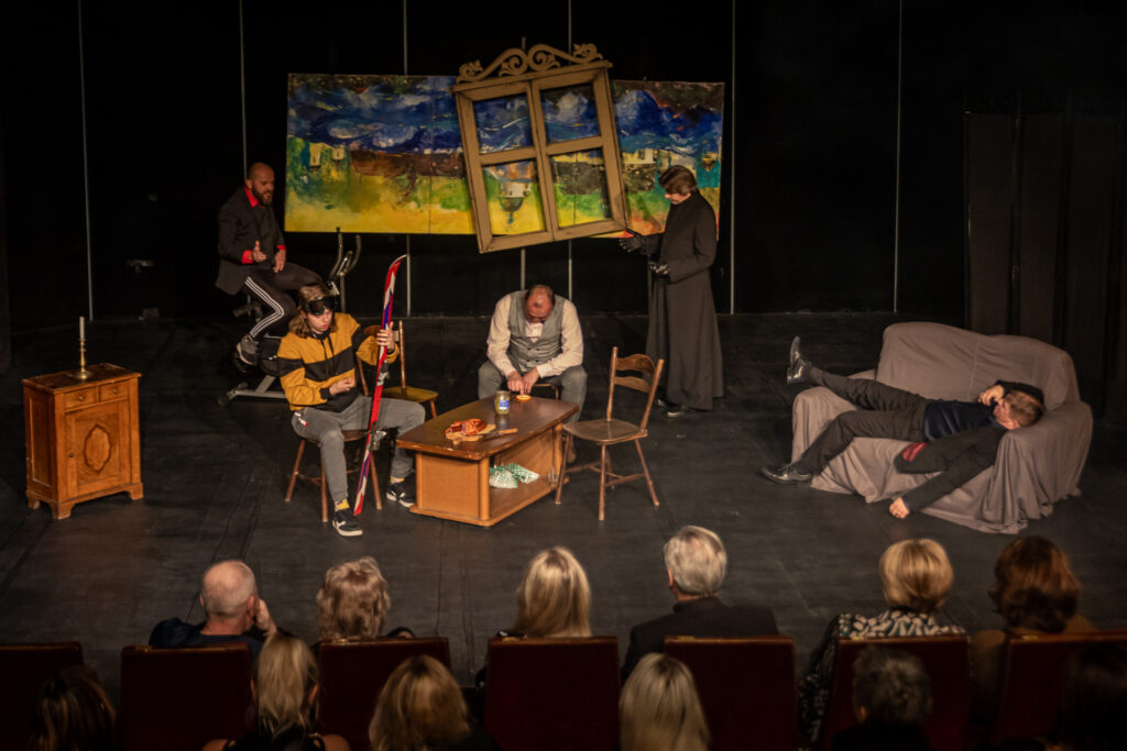 Zdjęcie. Mała Scena KCK, na niej aktorzy teatru Ciut Frapującego z Pińczowa. Młody chłopak smaruje narty, mężczyzna w kamizelce siedzi z opuszczoną głową, mężczyzna w ciemnym garniturze leży na kanapie, W tle stoi ksiądz i mężczyzna w dresie na stacjonarnym rowerze treningowym.