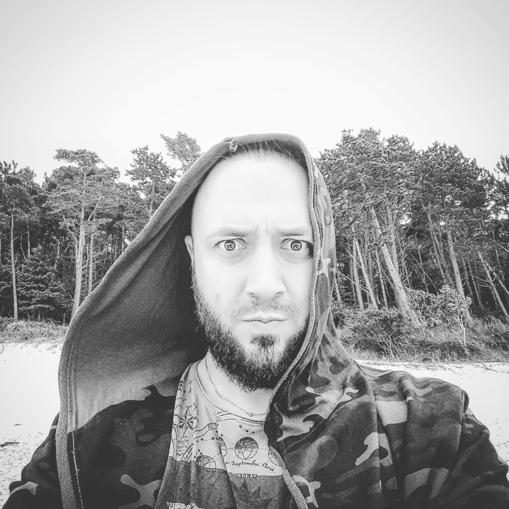 Czarno-białe zdjęcie. Krótko obcięty mężczyzna z brodą w kapturze bluzy na głowie z przestraszoną miną patrzy w obiektyw aparatu. To Grzegorz Bukowski, muzyk.