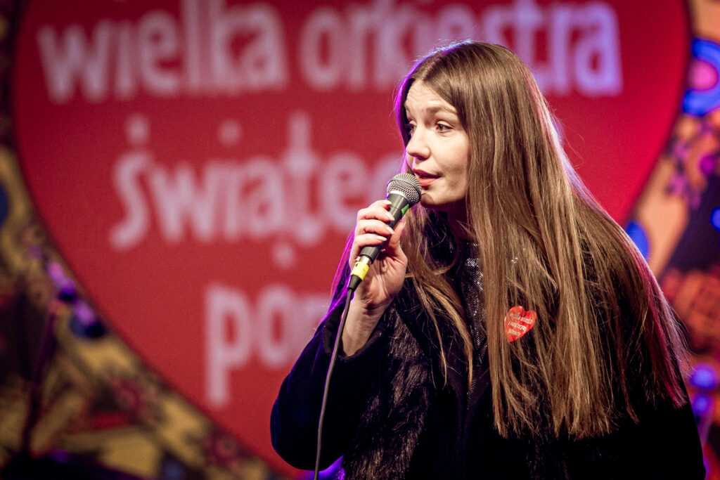 Zdjęcie. Długowłos blondynka o prostych rozpuszczonych włosach śpiewa do mikrofonu. To dyrektor KCK Agata Klimczak-Kołakowska.