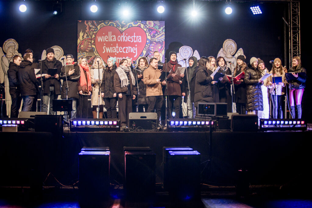 Zdjęcie. Duża grupa ludzi: konbiet i mężczyzn stoi na scenie i śpiewa do mikrofonów.