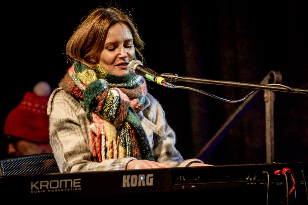 Zdjęcie. Kobieta o prostych, miodowych włosach, opatulona szerokim zielono-pomarańczowym szalem. Śpiewa domikrofonu, grając na keyboardzie. To Kasia Lisowska.