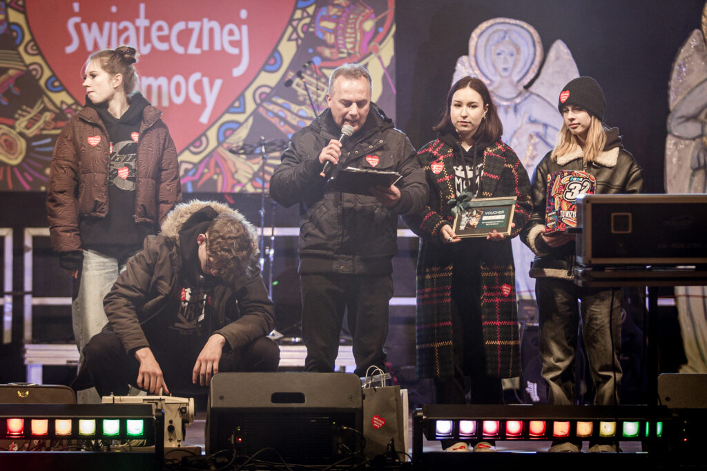 Zdjęcie. Prowadzący koncert finałowy mężczyzna mówi do mikrofonu, patrząc w kartki. Z tyłu wolontariuszki prezentują tabliczkę.
