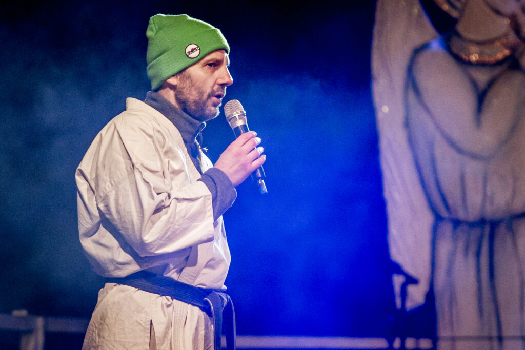 Zdjęcie. Karateka w kimonie i zielonej czapce na głowie mówi do mikrofonu.