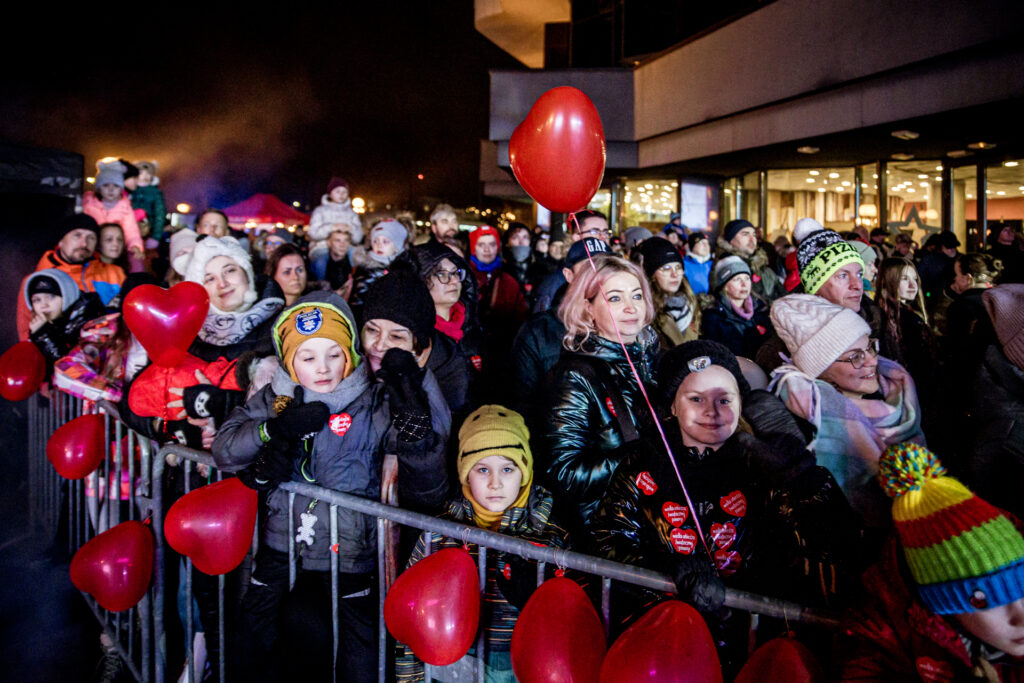 Zdjęcie. Widownia finału WOŚP na plenerowej scenie pod KCK. Dzieci i dorośli patrzą w kierunku sceny, stojąc za metalowymi bramkami, na których powieszono balony w kształcie serca.