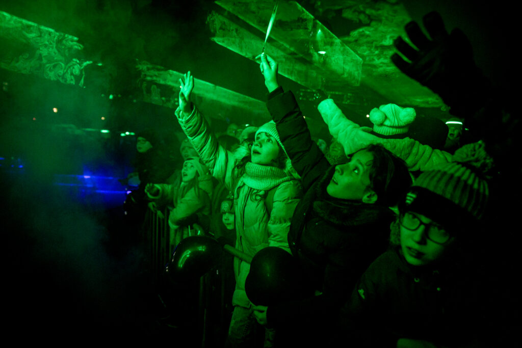 Zdjęcie. Widownia pokazu Światełko do Nieba podczas finału WOŚP. Ludzie wyciągają ręce do góry w kierunku zielonego światła laserowego.