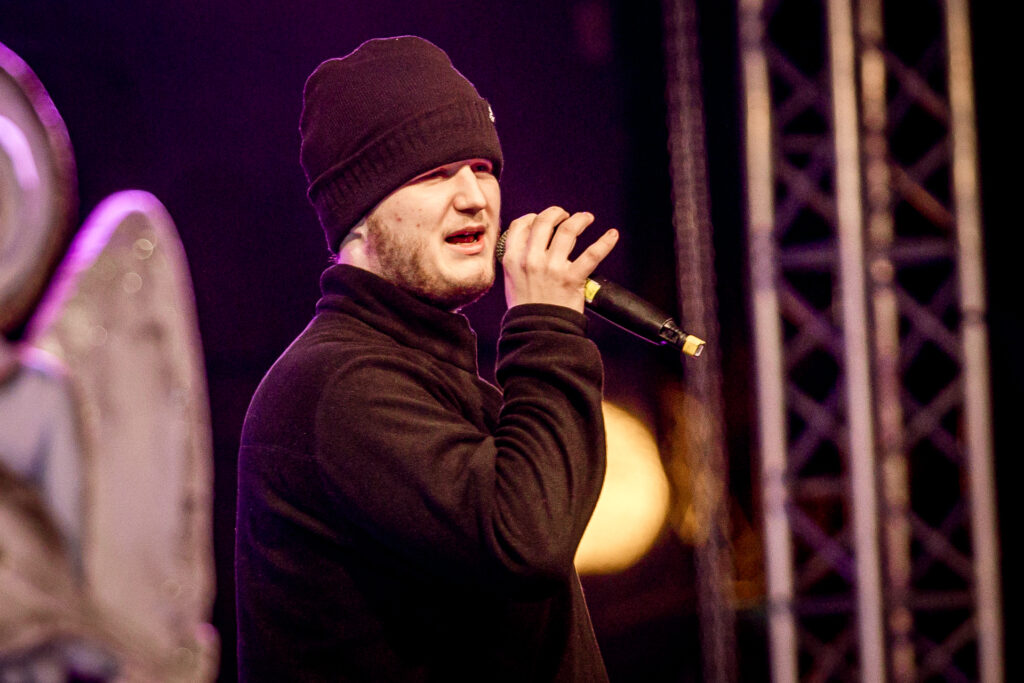 Zdjęcie. Młody mężczyzna z krótką brodą w czarnej czapce i bluzie mówi do mikrofonu.