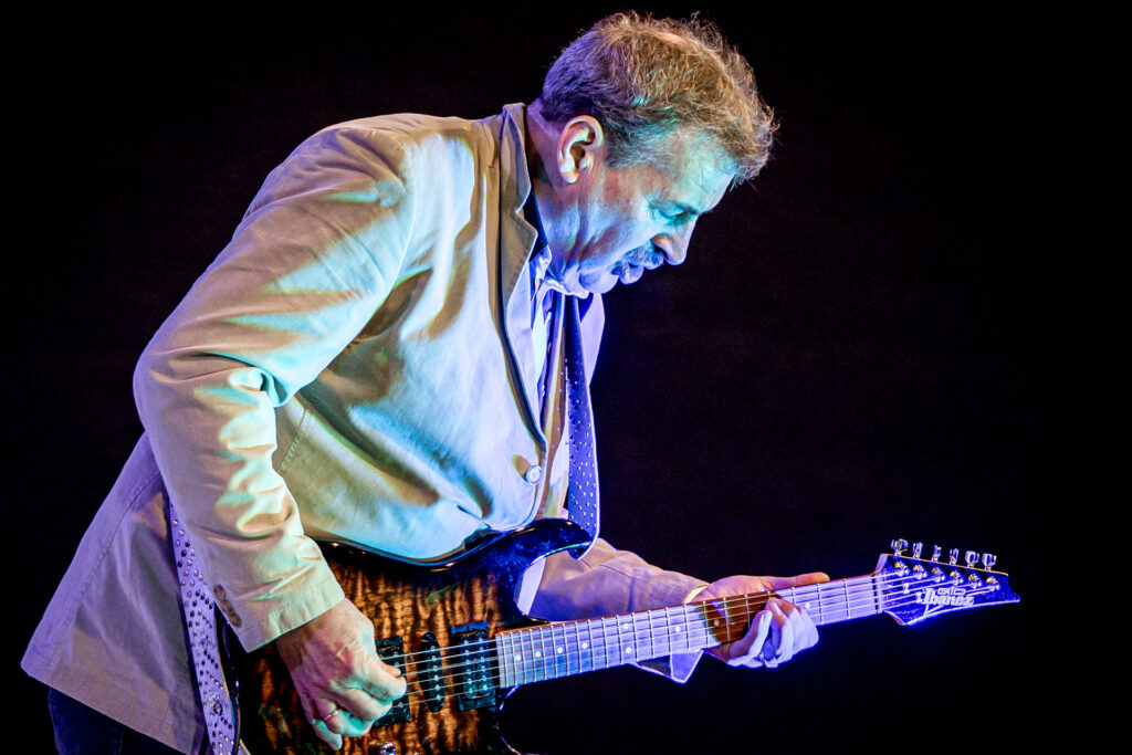 Zdjęcie. Starszy pan z wąsami w jasnej marynarce gra na gitarze.
