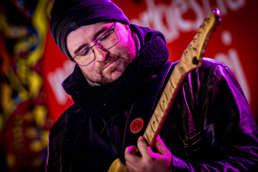 Zdjęcie. Mężczyzna z brodą w czarnej czapce i okularach gra na gitarze.