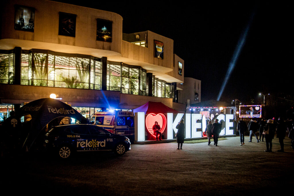Zdjęcie. Noc. Budynek KCK z oświetlonym Foyer. Przed budynkiem samochód z napisem: Radio Rekord, świecący napis: I love Kielce. W tle scena plenerowa. w nocy.
