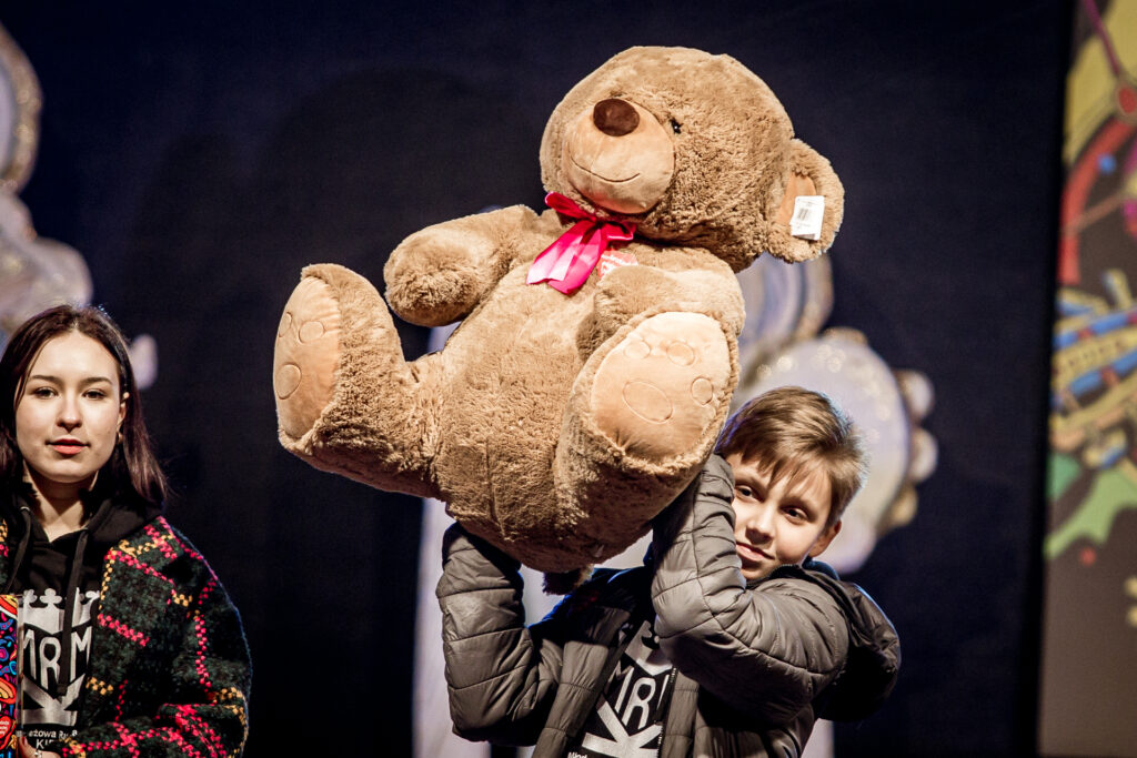 Zdjęcie. Mały chłopiec podnosi na scenie dużego pluszowego misia z czerwoną kokardą.