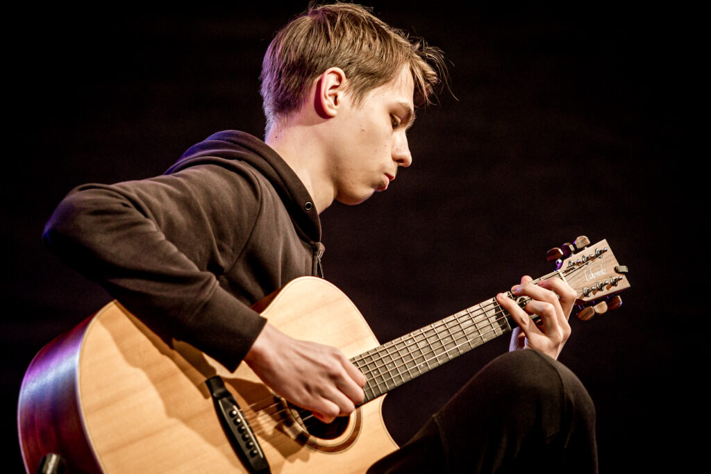 Zdjęcie. Młodzieniec w czarnej bluzie gra na gitarze.
