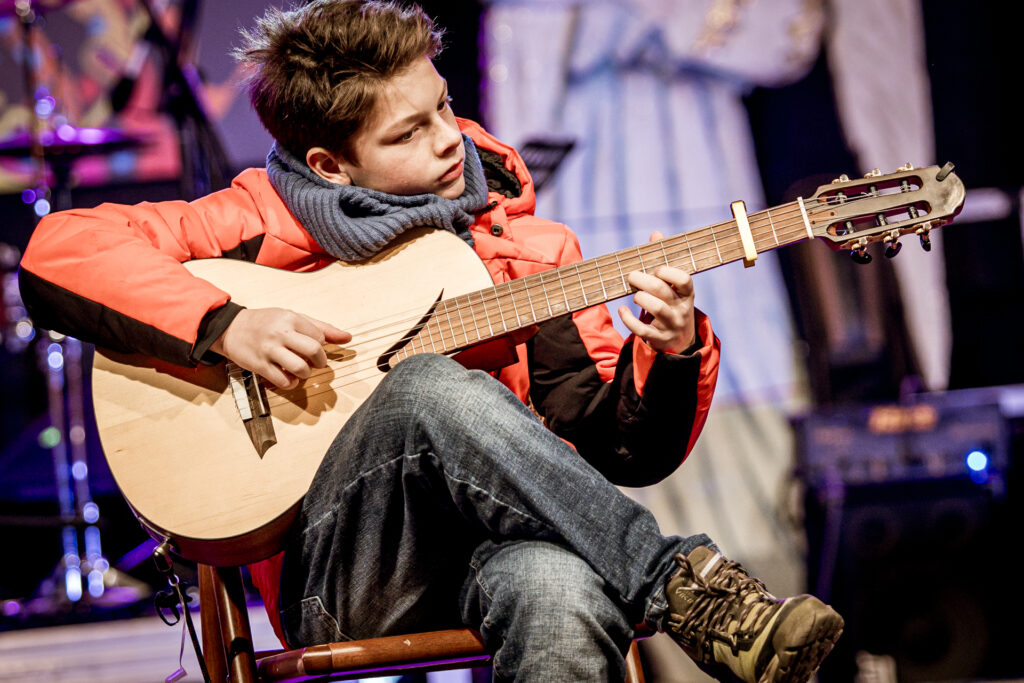 Zdjęcie. Młody chłopiec w czerwonej kurce siedzi na krześle na scenie i gra na gitarze.