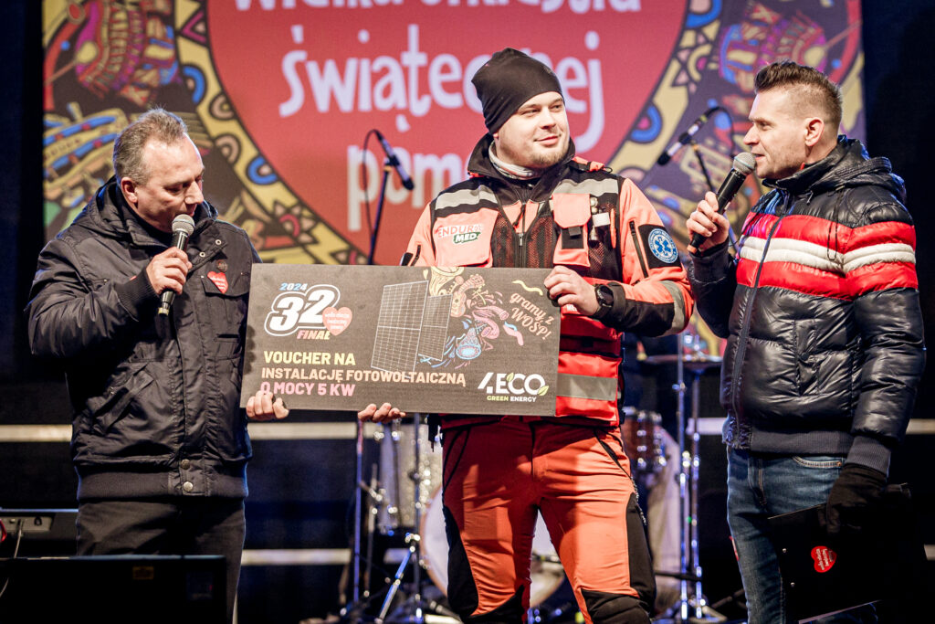Zdjęcie. Dwójka prowadzących na scenie plenerowej KCK. W środku stoi mężczyzna w stroju ratownika medycznego. W rękach trzyma dużą tavliczkę z napisem: voucher na instalację fotowoltaiczną o mocy 5 kW.