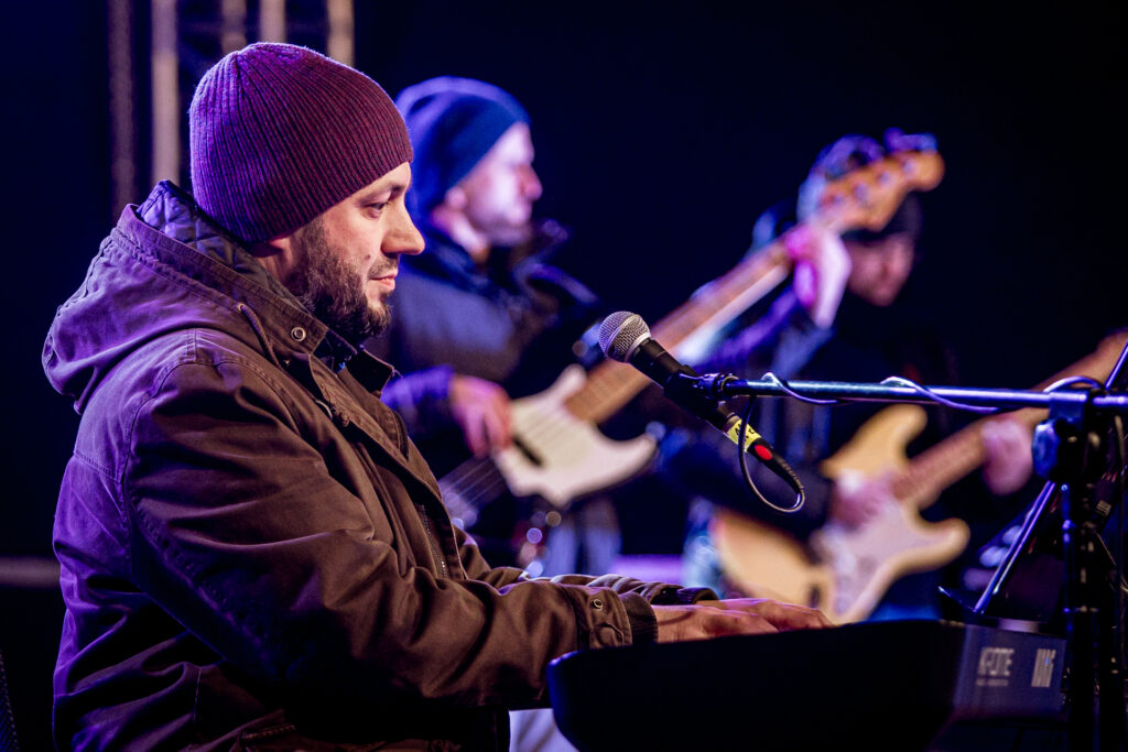 Zdjęcie. Mężczyzna z brodą w kurtce gra na keyboardzie. W tle ubrany w kurtkę gitarzysta. To finał WoŚP pod KCK.