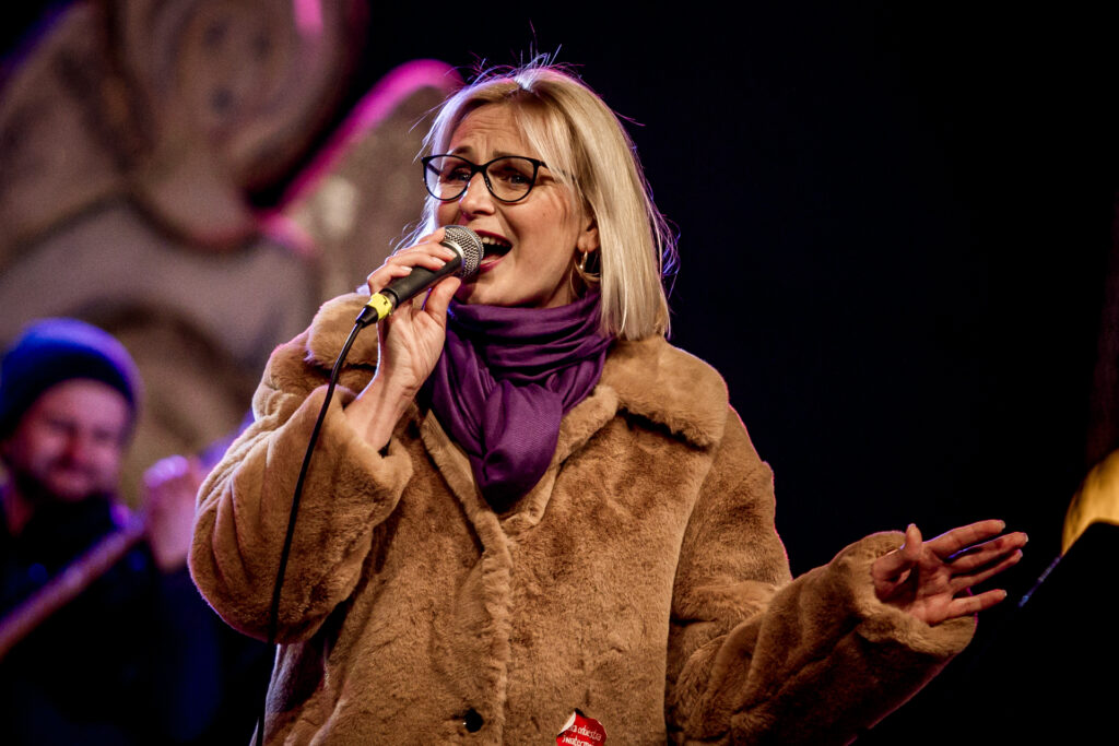 Zdjęcie. Blondynka z prostymi włosami w okularach w ciemnych oprawkach śpiewa do mikrofonu. To dyrektor DK Zameczek, Magdalena Nowaczek.