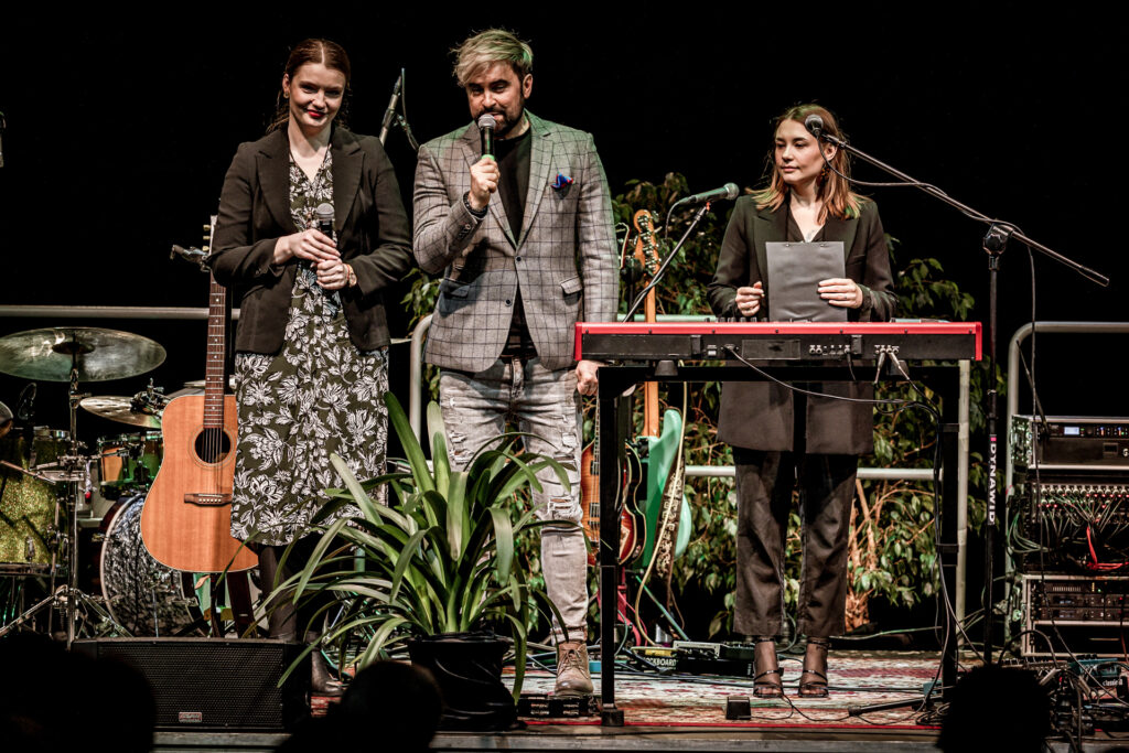 Zdjęcie. Koncert Podsłuchy. Wydarzenie zapowiada trójka ludzi. Od lewej uśmiechająca się dyrektor KCK Agata K.limczak-Kołakowska. Domikrofonu mówi uśmiechniety mężczyzna w szarej marynarce w czarną kratę. To wicedyrektor KCK Artur Wijata, ZA keyboardem stoi młoda dziewczyna ubrana w czarny garnitur. To pomysłodawczyni i koordynatorka Podsłuchów Daria Pol.