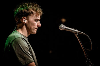 Zdjęcie. Koncert Podsłuchy. Patrick the Pan stoi z zamkniętymi oczami przed mikrofonem na statywie.