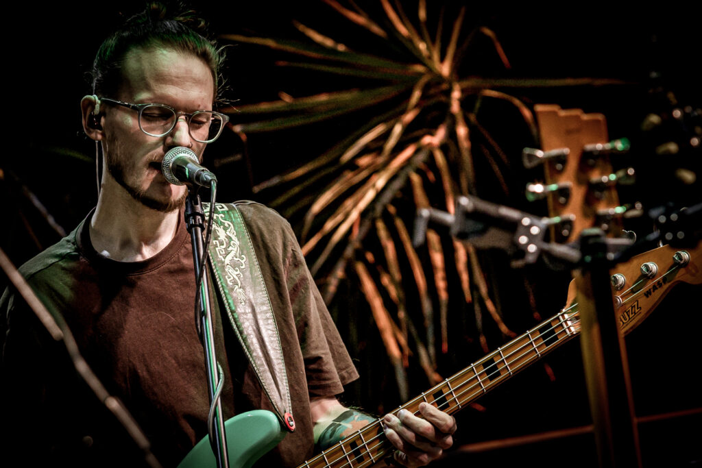 Zdjęcie. Koncert Podsłuchy. Mężcyznza w brązowej podkoszulcew i okularach gra na gitarze i śpiewa do mikrofonu. To gitarzysta grupy Patrick the Pan.