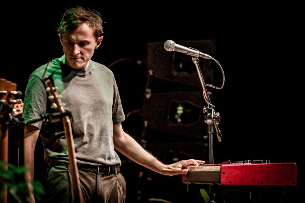 Zdjęcie. Koncert Podsłuchy. Mężczyzna opiera się lewą ręką o czerwony keyboard. Patrzy w prawą stronę w dół. To Patrick the Pan.