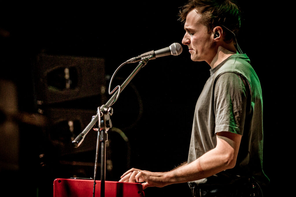 Zdjęcie. Koncert Podsłuchy. Mężczyzna w ciemnobeżowej podkoszulce stoi przy czerwonym keyboardzie. Przed sobą ma mikrofon na statywie. To Patrick the Pan.