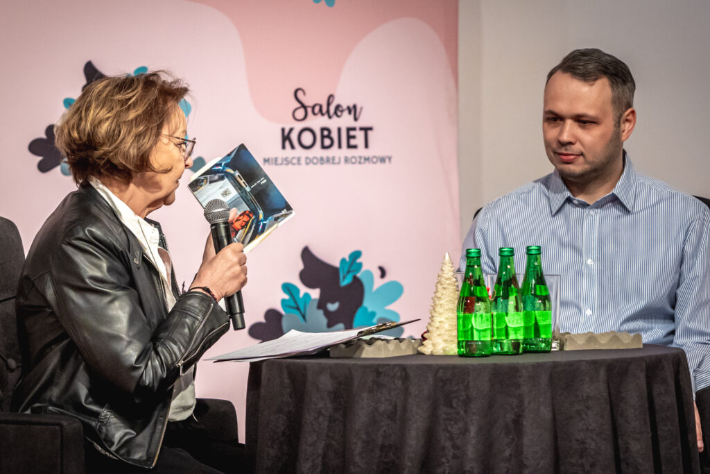 Zdjęcie. Dwie osoby: kobieta i mężczyzna rozmawiają przy stoliku . To Marzena Sobala, prowadząca Salon Kobiet i jej gość, lekarz Jakub Sieczko. Marzena Sobala mówi do mikrofonu. W lewej ręce unosi książkę, której autorem jest Jakub Sieczko.