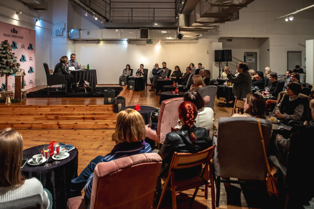 Zdjęcie. Widownia Salonu Kobiet w Galerii winda. Na podwyższeniu przy stoliku siedzi prowadząca i jej gość. Poniżej w fotelach przy stolikach - widzowie.