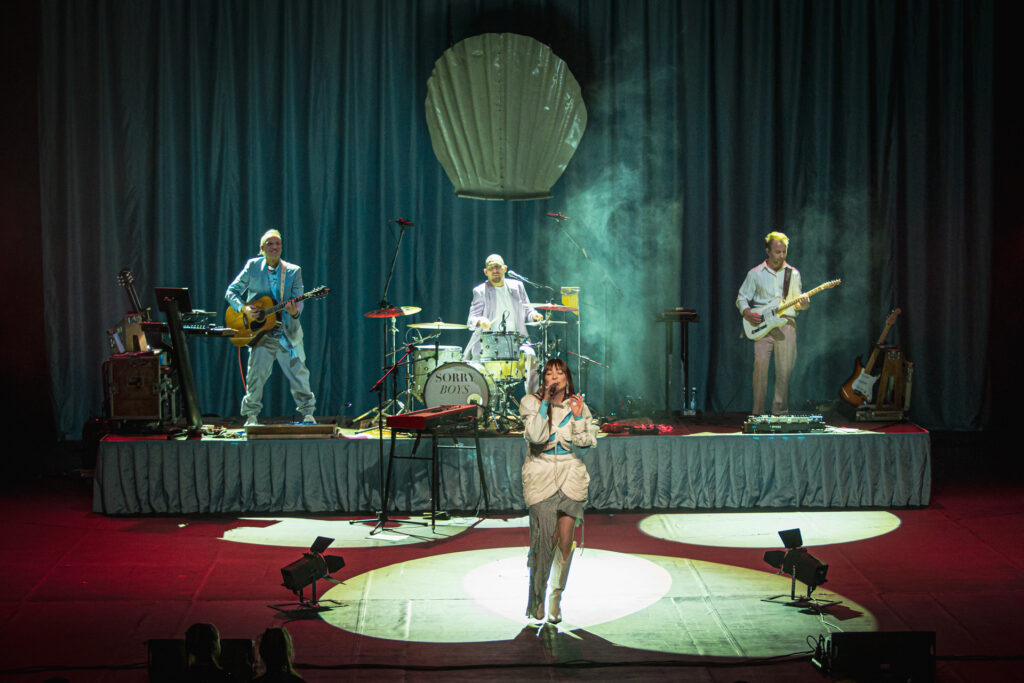 Zdjęcie. Duża Scena KCK podczas koncertu Sorry Boys. Z przodu wokalistka - Bela Komoszyńska. Z tyłu na podwyższeniu dwóch gitarzystów i perkusista. Nad nimi wielka plastikowa muszla.