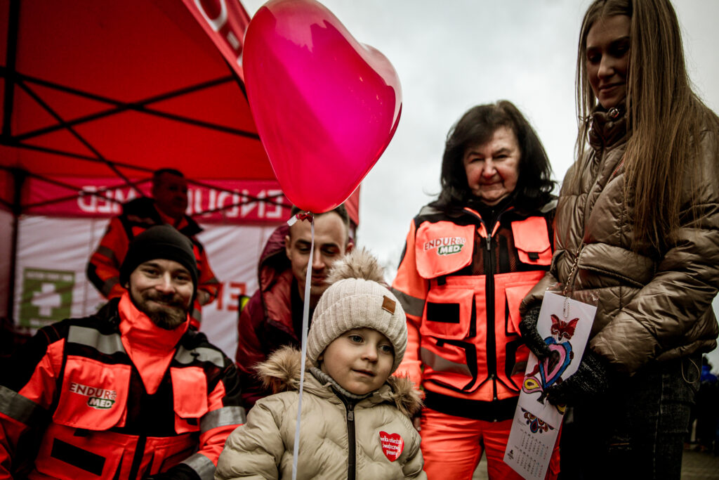 Zdjęcie. Dziewczynka wbeżowej kurtce i czapce z balonem w kształcie czerwonego serca pozuje z ratownikami medycznymi z grupy enduro -med. Obok stoi jej mama: blondynka o długich, rozpuszczonych prostych włosach.