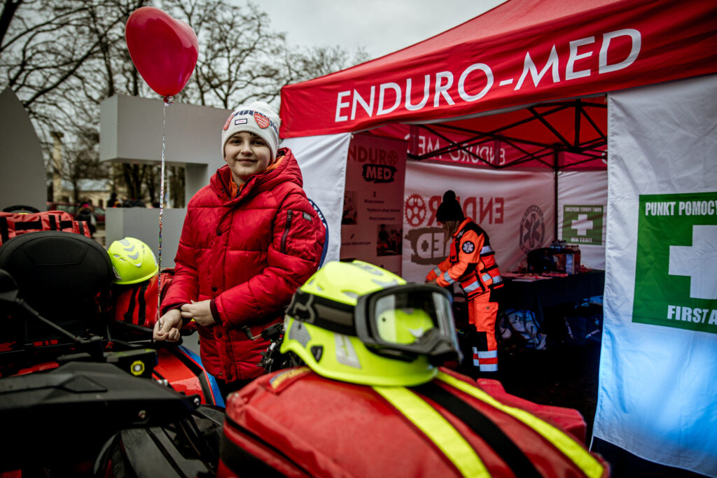 Zdjęcie. Chłopak w czerwonej kurtce i beżowej czapce pozuje w pobliżu namiotu z napisem enduro-med. W namiocie stoi mężczyzna w odblaskowym uniformie ratownika medycznego.