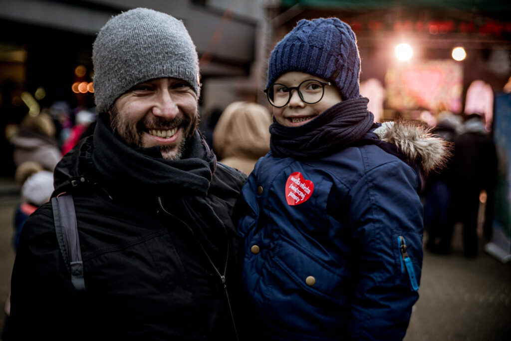 Zdjęcie. Uśmiechnięty mężczyzna z brodą trzyma na ręce kilkuletniego chłopca w granatowej kurtce, czapce i w okularach o czarnych oprawkach.