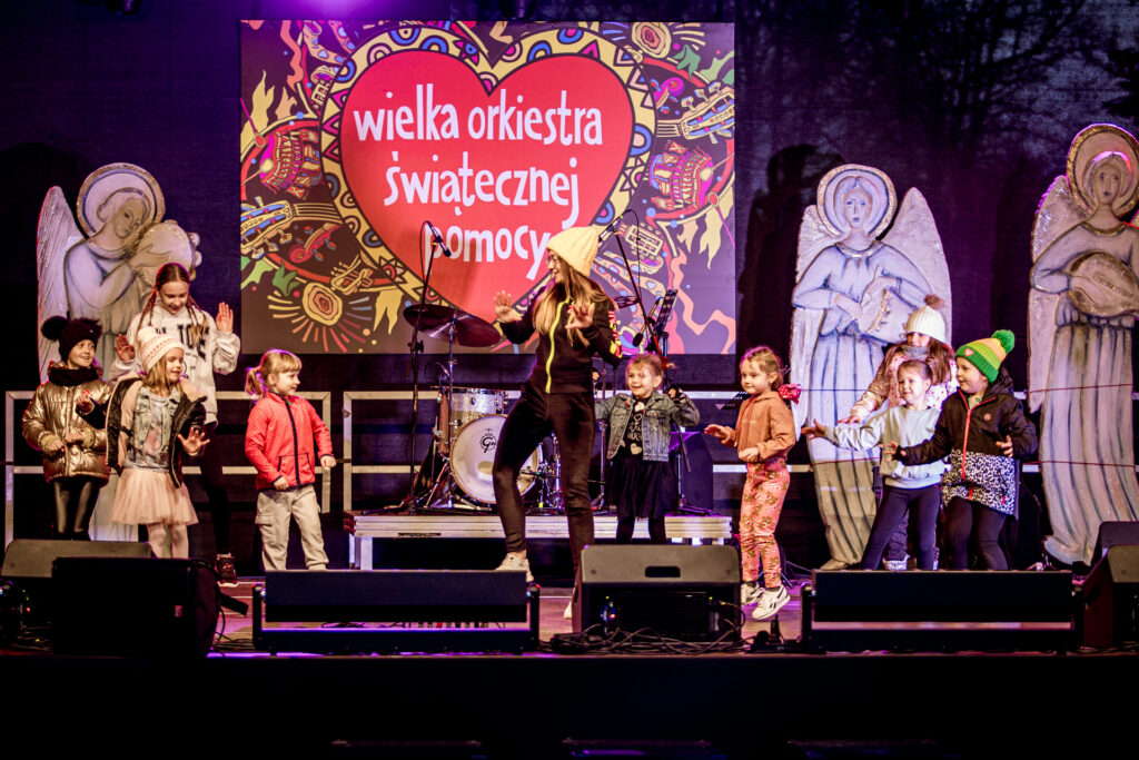 Zdjęcie. Młoda dziewczyna w czarnym dresie i białej czapce tańczy na środku sceny. Dookoła niej małe dzieci, które próbują ją naśladować.