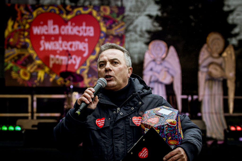Zdjęcie. Siwiejący mężczyzna mówi do mikrofonu. Pod pachą trzyma puszkę finału WOŚP. W tle plenerowa scena KCK z dwoma dużymi aniołami.