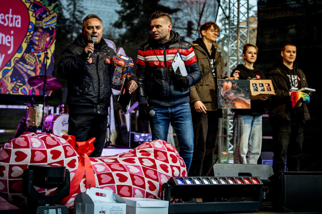 Zdjęcie. Dwójka prowadzących mówi do mikrofonu. W tle trójka wolontariuszy trzymających fanty do licytacji.