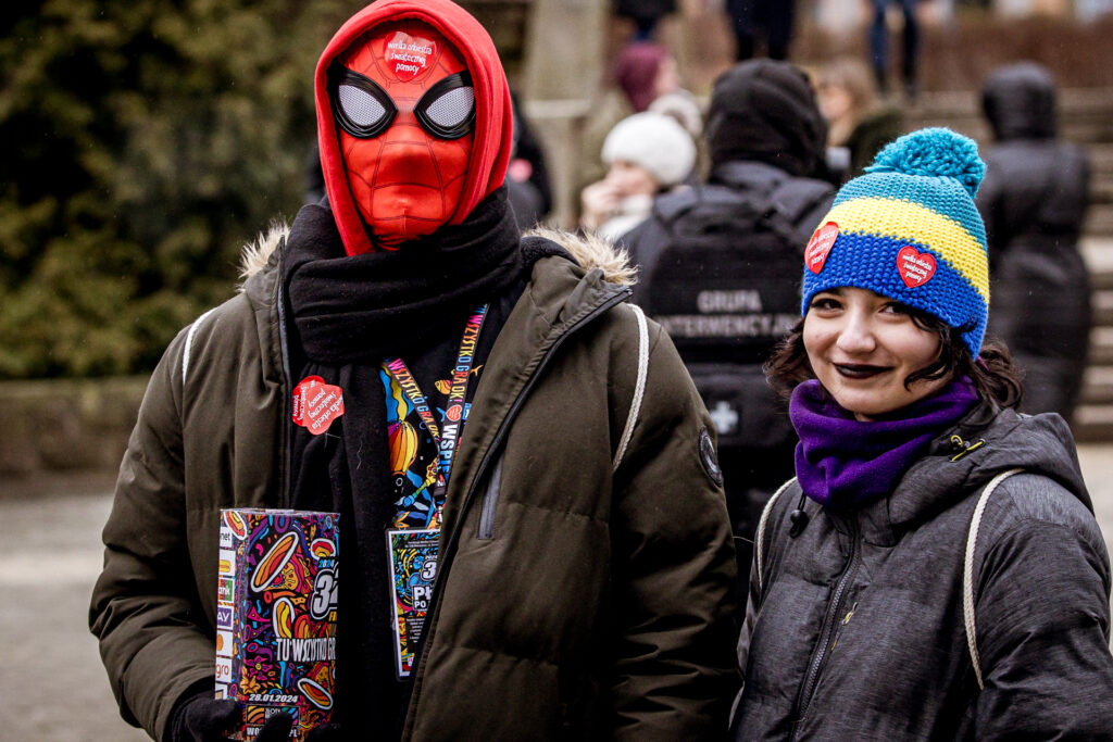 Zdjęcie. Dwójka wolontariuszy WOŚP: Chłopak i dziewczyna z ustami pomalowanymi na czarno pozują fotografowi. Chłopak ma na twarzy czerwoną maskę Spidermana.