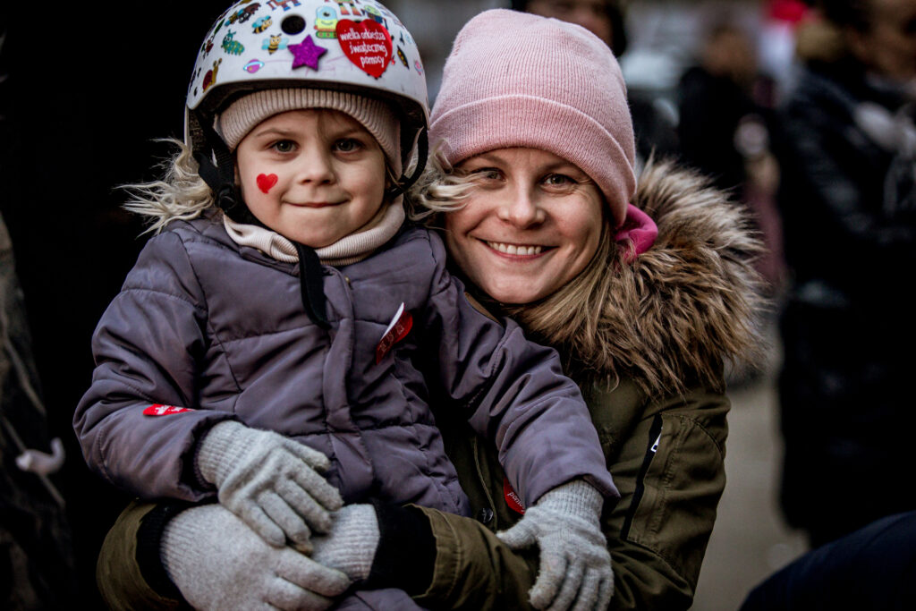 Zdjęcie. Kobieta trzyma na kolanie i obejmuje w pasie małą dziewczynkę w białym kasku na głowie. Obie uśmiechają się do obiektywu.