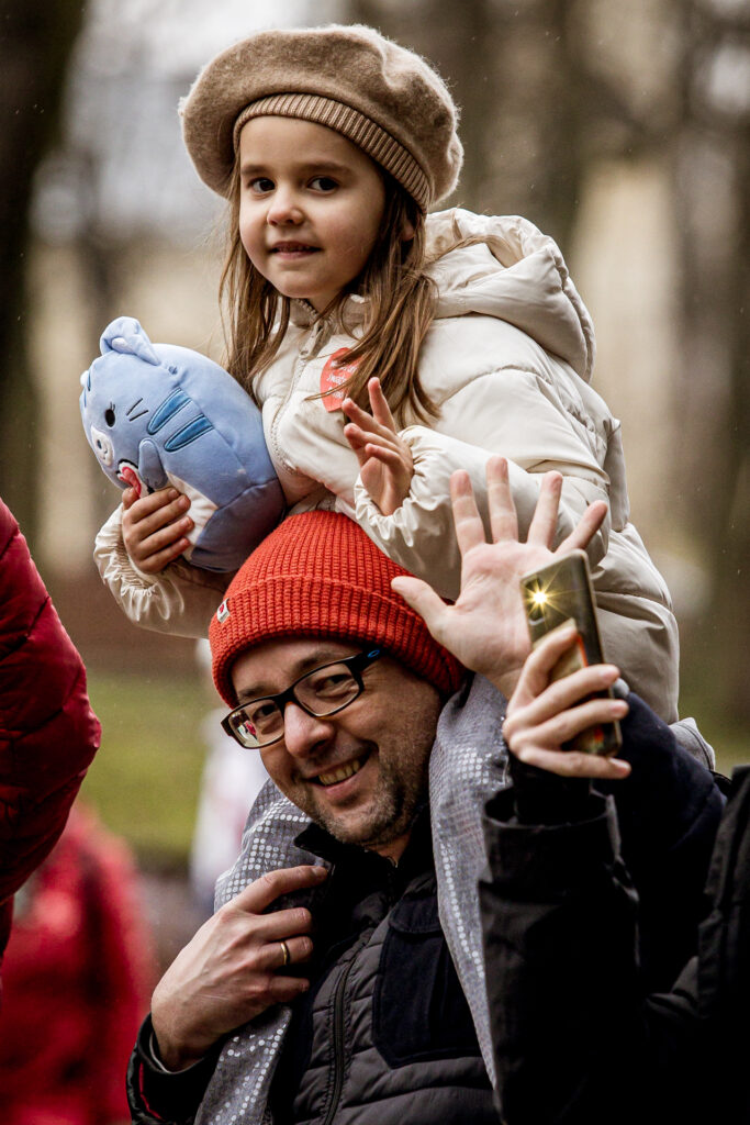 Zdjęcie. Dziewczyna w beżowej kurtce i berecie siedzi na "barana" na ramionach mężczyzny w czerwnej czapce i okularach. Oboje machają do fotografa. To widzowie finału WOŚP pod KCK.