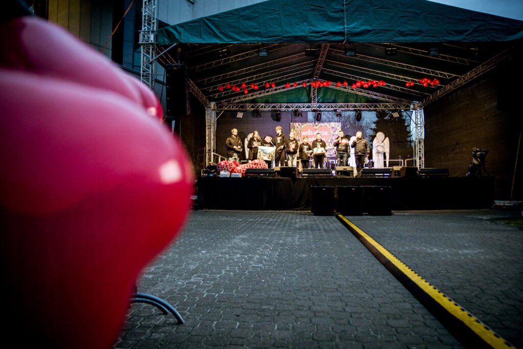 Zdjęcie. Duża plenerowa scena KCK podvzas dinału WOŚP. Na scenie stoi rząd ludzi z przedmiotami do licytacji w dłoniach.