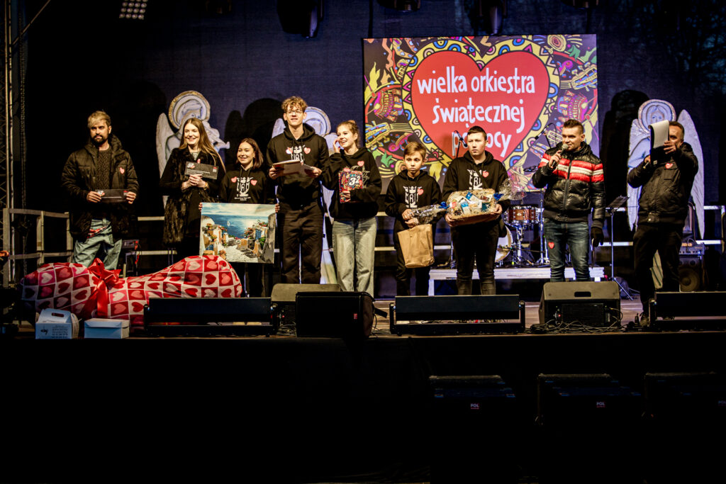 Zdjęcie. Dziewięć osób na scenie plenerowej finału Wość. Od lewej: wicedyrketor KCK Artur Wijata, dyrektor KCK Agata Klimczak-Kołakowska, piątka dzieci i młodzieży - wolontariuszy WOŚP oraz dwójka prowadzących. Osoby poza prowadząctcmi trzymają w rękach przedmioty do licytacji.