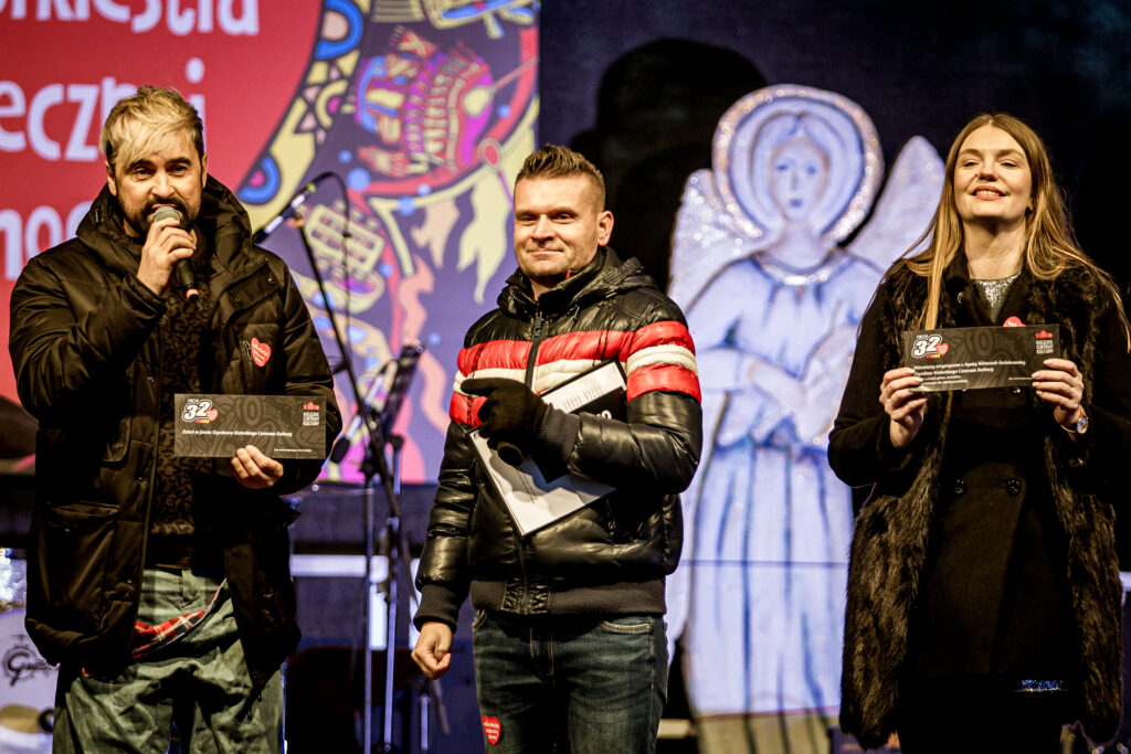 Zdjęcie. Trójka ludzi na scenie. Od lewej: wicedyrektor Artur Wijata mówi do mikrofonu. Uśmiechnięty prowadzący koncert. Obok stoi uśmiechnięta dyrektor KCK Agata Klimczak-Kołakowska.