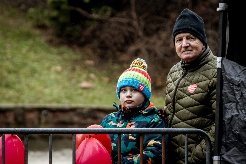Zdjęcie. Starszy mężczyzna w ciemnej czapce i ciemnozielonej kurtce, a przed nim chłopiec w kolorowej czapce i kurtce patrzą w kierunku obiektywu. To widzowie finału WOŚP przed KCK.