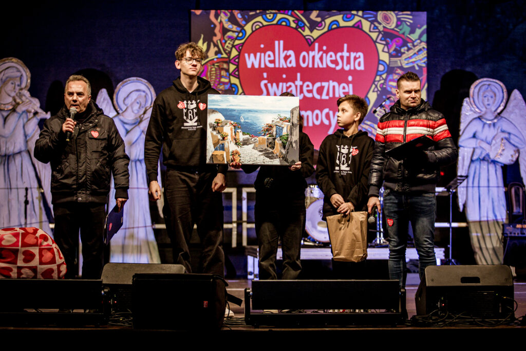 Zdjęcie. Plenerowa scena finału WOŚP. Na niej wójka wolontariuszy pokazuje duży obraz lub zdjęcie z widokiem morza i położonych na wyboirzeżu domów w klimacie południa Europy. Obok wolontariuszy stoją dwaj prowadzący. Jeden z nich mówi do mikrofonu.