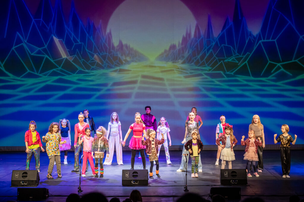 Zdjęcie. Duża grupa dzieci stoi na scenie w kilku rzędach w pewnej odległości od siebie. W tle kolorowa wizualizacja. To pokaz musicalu MOC.