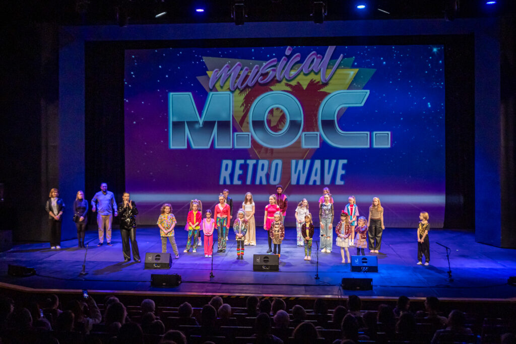 Zdjęcie. Grupa dzieciaków stoi na scenie. W tle napis: musical Moc Retro Wave. Obok instruktorzy warsztatów ferie z musicalem. 
