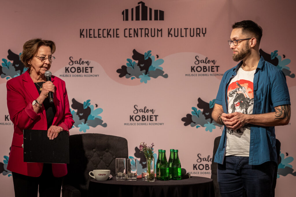 Zdjęcie. DWójka ludzi stoi przy okrągłym stoliku. Kobieta w okularach i czerwonej marynarce i mody mężczyzna w białej podkoszulce z nadrukiem i niebieskiej koszuli z krótkim rękawem. W tle różowa ścianka z napisami Salon Kobiet.