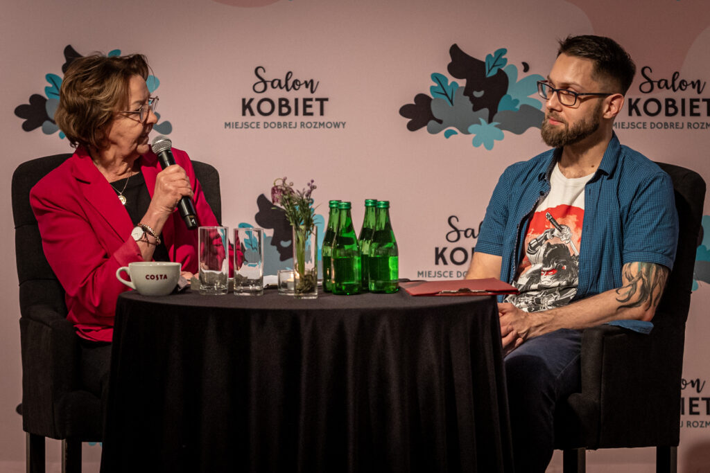 Zdjęcie. DWójka ludzi siedzi przy okrągłym stoliku. Kobieta w okularach i czerwonej marynarce i mody mężczyzna w białej podkoszulce z nadrukiem i niebieskiej koszuli z krótkim rękawem. W tle różowa ścianka z napisami Salon Kobiet.