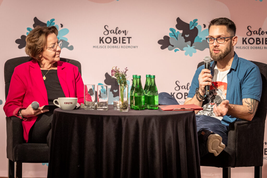 Zdjęcie. DWójka ludzi siedzi przy okrągłym stoliku. Kobieta w okularach i czerwonej marynarce i mody mężczyzna w białej podkoszulce z nadrukiem i niebieskiej koszuli z krótkim rękawem. W tle różowa ścianka z napisami Salon Kobiet.