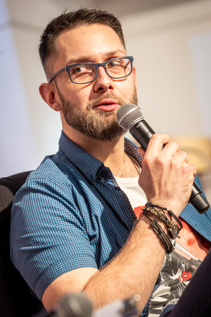 Zdjęcie. Młody mężczyzna w białej podkoszulce z nadrukiem i niebieskiej koszuli z krótkim rękawem. mówi do mikrofonu. To Maciej Frasunkiewicz. Ma krótko obcięte włosy, brodę oraz okulary w ciemnych oprawkach.