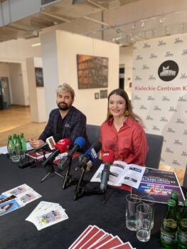 Zdjęcie. Dwójka ludzi siedzi za dużym stołem. To dyrektor KCK Agata Klimczak-Kołakowska w pomarańczowej koszuli. Kobieta uśmiecha się. Obok mężczyzna jasną grzywką i ciemną brodą i wąsami wicedyrektor Artur Wijata. Na stole plakaty, ulotki oraz mikrofony stacji radiowych i telewizyjnych.